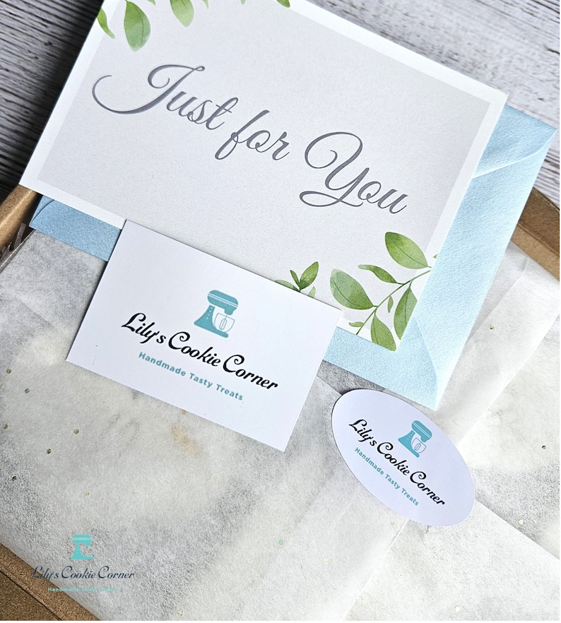 Pode incluir: Uma caixa de presente com um cart&atilde;o que diz "Just for You" em uma escrita elegante, adornado com ilustra&ccedil;&otilde;es de folhas verdes. A caixa inclui um envelope azul claro e cart&otilde;es de visita com o logotipo "Lily's Cookie Corner" e o texto "Handmade Tasty Treats."