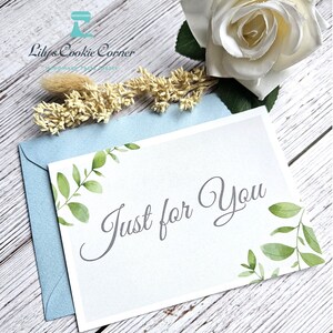 Pode incluir: Um cart&atilde;o branco com o texto "Just for You" em escrita elegante, adornado com folhas verdes em aquarela. Repousa sobre um envelope azul claro, com uma rosa branca e detalhes florais secos. Um gesto atencioso.