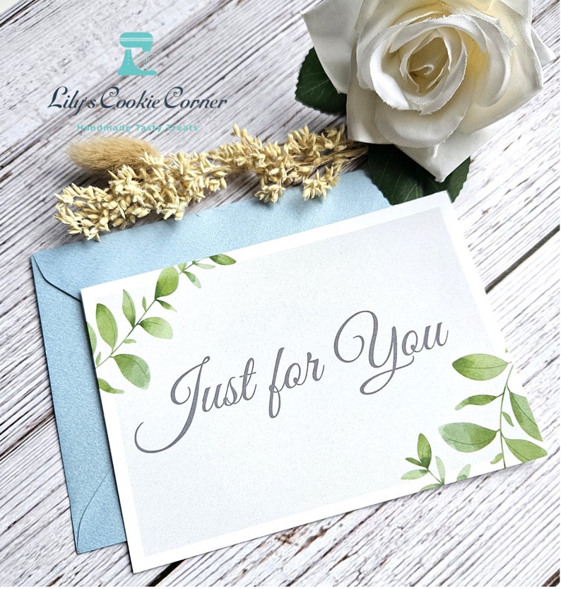 Pode incluir: Um cart&atilde;o branco com o texto "Just for You" em escrita elegante, adornado com folhas verdes em aquarela. Repousa sobre um envelope azul claro, com uma rosa branca e detalhes florais secos. Um gesto atencioso.