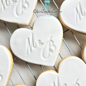 May include: Heart-shaped cookies with white icing and the initials "M & B" embossed on top. The cookies are arranged on a metal cooling rack. The cookies have a golden-brown edge. The image also includes the text "Lily's Cookie Corner" and "Handmade Tasty Treats".