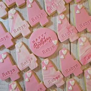 May include: A collection of pink and white cookies, some shaped like the number four, others round. The cookies are decorated with the name "Robyn", hearts, and the words "Happy Birthday". The cookies are arranged on a light wooden surface.