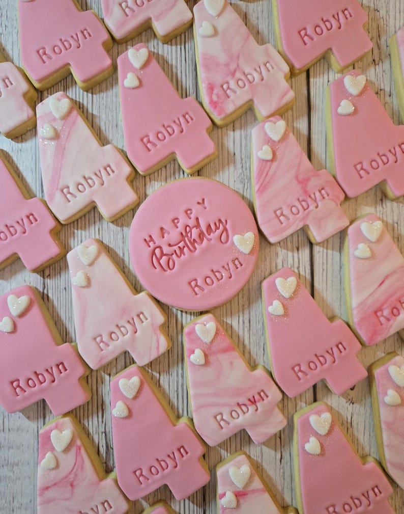 May include: A collection of pink and white cookies, some shaped like the number four, others round. The cookies are decorated with the name "Robyn", hearts, and the words "Happy Birthday". The cookies are arranged on a light wooden surface.