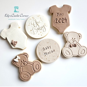May include: Five decorated sugar cookies for a baby shower. Two cookies are shaped like onesies, one is a circle with the text "Oh Baby", one is a circle with the text "Baby Moran", and two are teddy bears.
