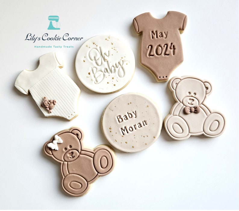 May include: Five decorated sugar cookies for a baby shower. Two cookies are shaped like onesies, one is a circle with the text "Oh Baby", one is a circle with the text "Baby Moran", and two are teddy bears.