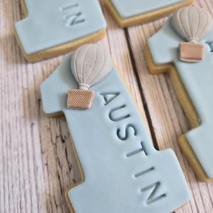 May include: Light blue number one cookies with the name "AUSTIN" imprinted on them. Each cookie has a light gray hot air balloon with a brown basket attached. The cookies are on a light wood surface.