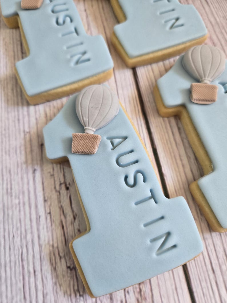 May include: Light blue number one cookies with the name "AUSTIN" imprinted on them. Each cookie has a light gray hot air balloon with a brown basket attached. The cookies are on a light wood surface.