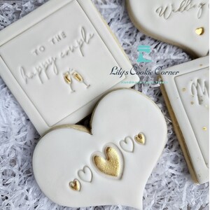 Pode incluir: Close-up de biscoitos de a&ccedil;&uacute;car decorados. Um biscoito quadrado diz "TO THE happy couple" com ta&ccedil;as de champanhe. Um biscoito em forma de cora&ccedil;&atilde;o tem cora&ccedil;&otilde;es dourados e brancos. Outros biscoitos dizem "Oh you Wedding" e "Mr & Mrs".