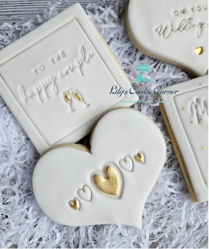 Pode incluir: Close-up de biscoitos de a&ccedil;&uacute;car decorados. Um biscoito quadrado diz "TO THE happy couple" com ta&ccedil;as de champanhe. Um biscoito em forma de cora&ccedil;&atilde;o tem cora&ccedil;&otilde;es dourados e brancos. Outros biscoitos dizem "Oh you Wedding" e "Mr & Mrs".
