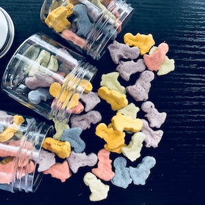 Puede incluir: Tres tarros de vidrio llenos de golosinas para perros de colores. Las golosinas tienen forma de hueso y son de tonos amarillos, naranjas, morados y azules. Los tarros se est&aacute;n derramando sobre una superficie negra.
