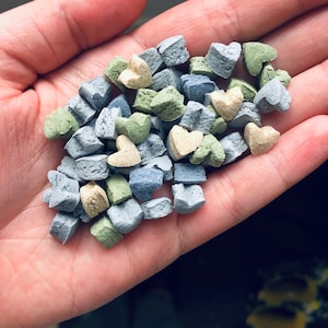 Puede incluir: Una colección de pequeños caramelos en forma de corazón en tonos azules, verdes y beige. Los caramelos están esparcidos en una mano.