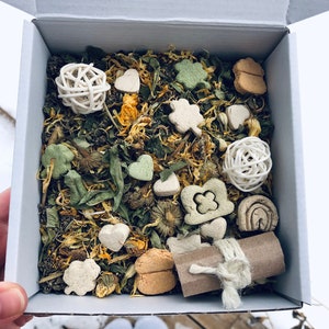 May include: A white cardboard box filled with a variety of dried herbs, flowers, and small, colourful, heart-shaped, clover-shaped, and other shaped treats for pets. The box is open and the contents are visible. There are two white wicker balls in the box.
