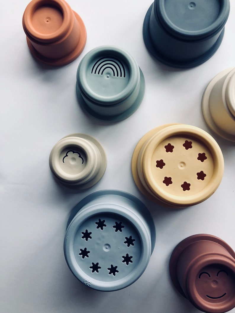 May include: Set of colorful stacking cups with different designs, including a rainbow, a cloud, stars, and a smiley face. The cups are made of silicone and are perfect for bath time or playtime.
