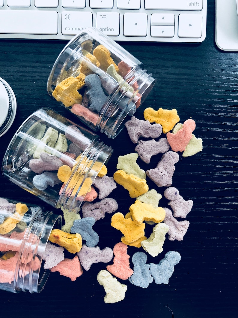 Puede incluir: Tres tarros de vidrio llenos de golosinas para perros de colores. Las golosinas tienen forma de hueso y son de tonos amarillos, naranjas, morados y azules. Los tarros se est&aacute;n derramando sobre una superficie negra.