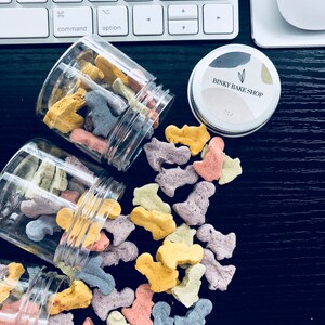 Puede incluir: Tres tarros de vidrio transparente llenos de golosinas para perros de colores. Las golosinas tienen forma de huesos peque&ntilde;os y son de tonos amarillos, naranjas, rosas, morados y grises. Los tarros est&aacute;n sobre una superficie negra con un teclado blanco y un tarro plateado con una etiqueta que dice "BINKY BAKE SHOP" en el fondo.
