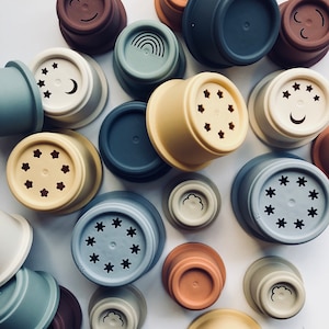 Può includere: Una collezione di tazze impilabili in silicone colorate con vari disegni, tra cui stelle, lune, nuvole, arcobaleni e facce sorridenti. Le tazze sono realizzate in silicone morbido e flessibile e sono perfette per impilare, annidare e giocare con i sensi.