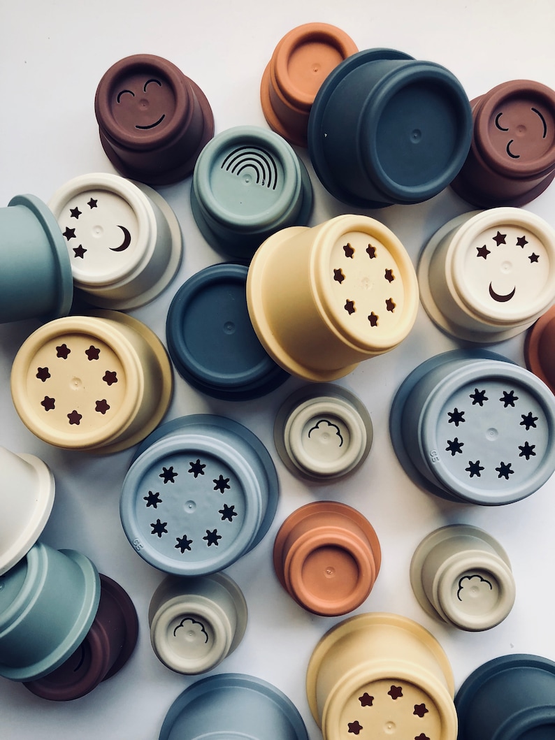 May include: A collection of colorful silicone stacking cups with various designs, including stars, moons, clouds, rainbows, and smiley faces. The cups are made of soft, flexible silicone and are perfect for stacking, nesting, and sensory play.