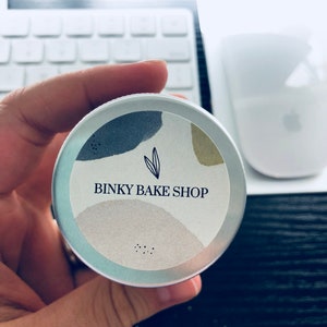 Puede incluir: Una lata de plata con una etiqueta blanca que dice "Binky Bake Shop" en letras negras. La etiqueta tiene un dise&ntilde;o abstracto azul claro, gris y beige con un peque&ntilde;o icono de hoja.