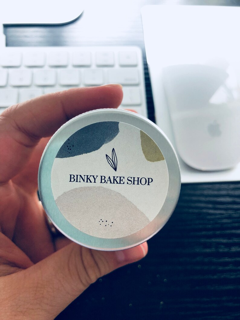 Puede incluir: Una lata de plata con una etiqueta blanca que dice "Binky Bake Shop" en letras negras. La etiqueta tiene un dise&ntilde;o abstracto azul claro, gris y beige con un peque&ntilde;o icono de hoja.