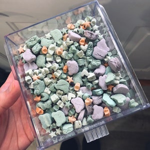 Könnte beinhalten: Ein durchsichtiger Plastikbehälter gefüllt mit pastellfarbenen Süßigkeiten. Die Süßigkeiten sind in Form von Sternen, Herzen und anderen geometrischen Formen. Die Süßigkeiten sind eine Mischung aus Grün, Blau, Rosa und Orange.