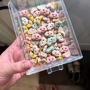 Könnte beinhalten: Ein durchsichtiger Plastikbehälter gefüllt mit buntem Müsli. Das Müsli ist in Form von Wolken und Monden mit Smiley-Gesichtern.