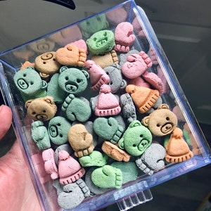 Könnte beinhalten: Ein durchsichtiger Plastikbehälter gefüllt mit bunten Bären- und Fäustlingsförmigen Keksen. Die Kekse sind in Grün-, Rosa-, Braun- und Grautönen.