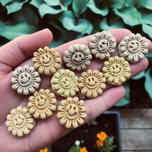 Könnte beinhalten: Eine Hand hält eine Sammlung kleiner, gelber und grüner Keramik-Blumenanhänger mit Smiley-Gesichtern. Die Anhänger sind in einem Halbkreis angeordnet.