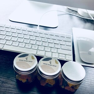 Puede incluir: Tres frascos de vidrio con tapas plateadas y contenido colorido. Cada frasco tiene una etiqueta blanca con el texto "Binky Bake Shop" en negro.