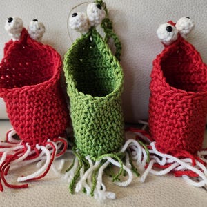 May include: Three handmade crochet monster-like hanging baskets. Two are red, and one is green. Each has white and red or green and white fringe at the bottom and white eyes. The baskets are likely for small items.
