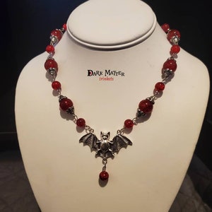 May include: A red beaded necklace with a silver bat pendant. The necklace is on a white mannequin bust. The text "DARK MATTER trinkets" is visible on the bust.