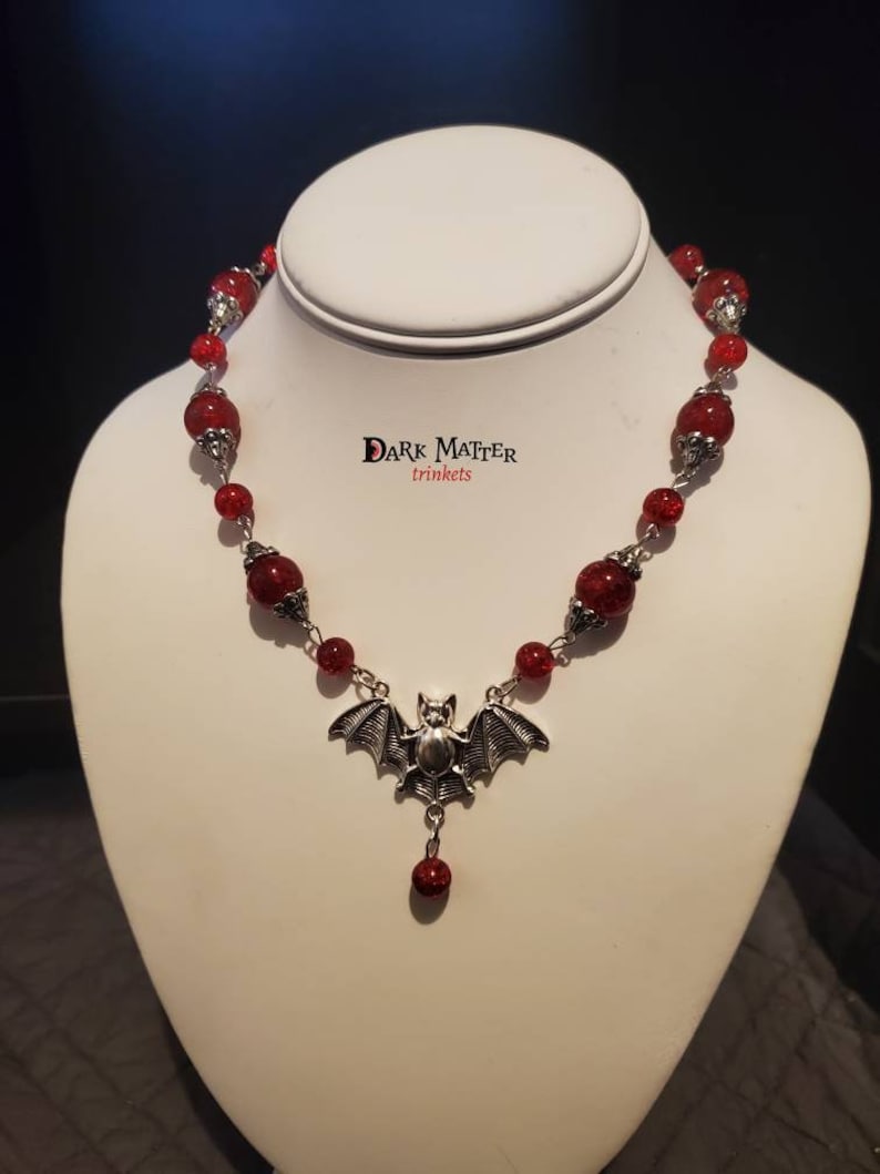 May include: A red beaded necklace with a silver bat pendant. The necklace is on a white mannequin bust. The text "DARK MATTER trinkets" is visible on the bust.