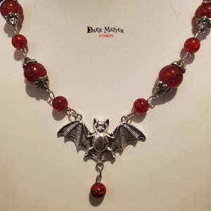 May include: A silver bat pendant necklace with a red glitter bead chain. The pendant features a bat with outstretched wings and a small red glitter bead hanging from the bottom. The necklace is labeled "Dark Matter Trinkets".