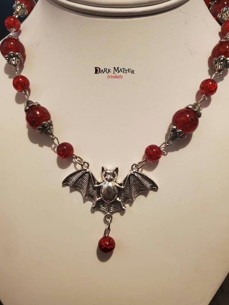 May include: A silver bat pendant necklace with a red glitter bead chain. The pendant features a bat with outstretched wings and a small red glitter bead hanging from the bottom. The necklace is labeled "Dark Matter Trinkets".