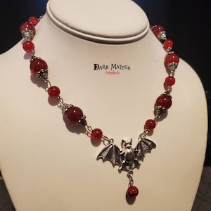 May include: A red glass bead necklace with a silver bat charm and a single red bead dangling from the bat. The necklace is on a white mannequin bust. The text "DARK MATTER trinkets" is visible on the bust.