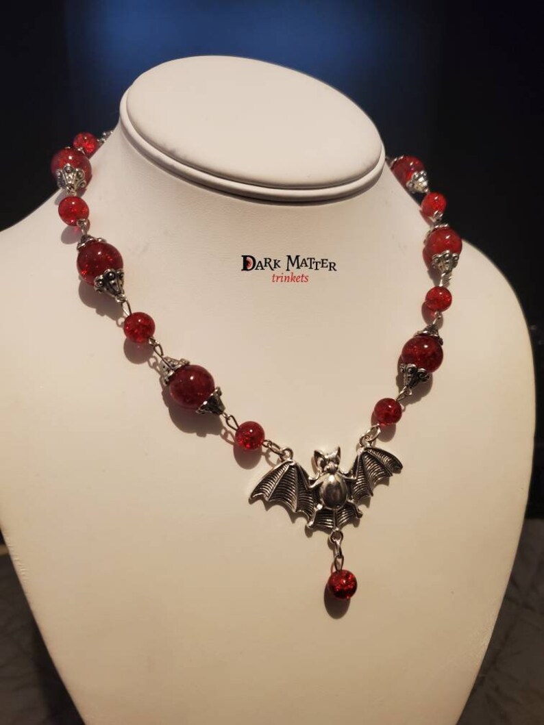 May include: A red glass bead necklace with a silver bat charm and a single red bead dangling from the bat. The necklace is on a white mannequin bust. The text "DARK MATTER trinkets" is visible on the bust.