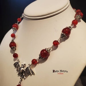 May include: A red beaded necklace with a silver bat pendant. The necklace is on a white mannequin bust. The text "DARK MATTER trinkets" is visible on the bust.
