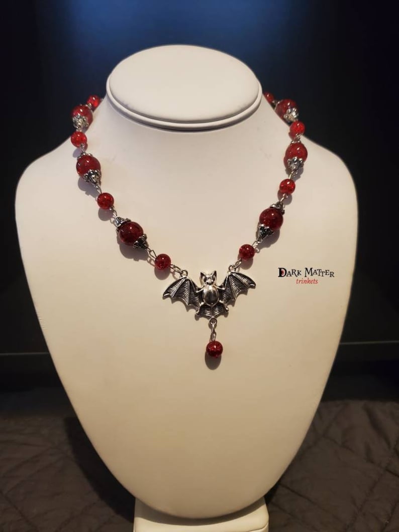 May include: A red beaded necklace with a silver bat pendant. The necklace is on a white mannequin bust. The text "DARK MATTER trinkets" is visible in the bottom right corner of the image.