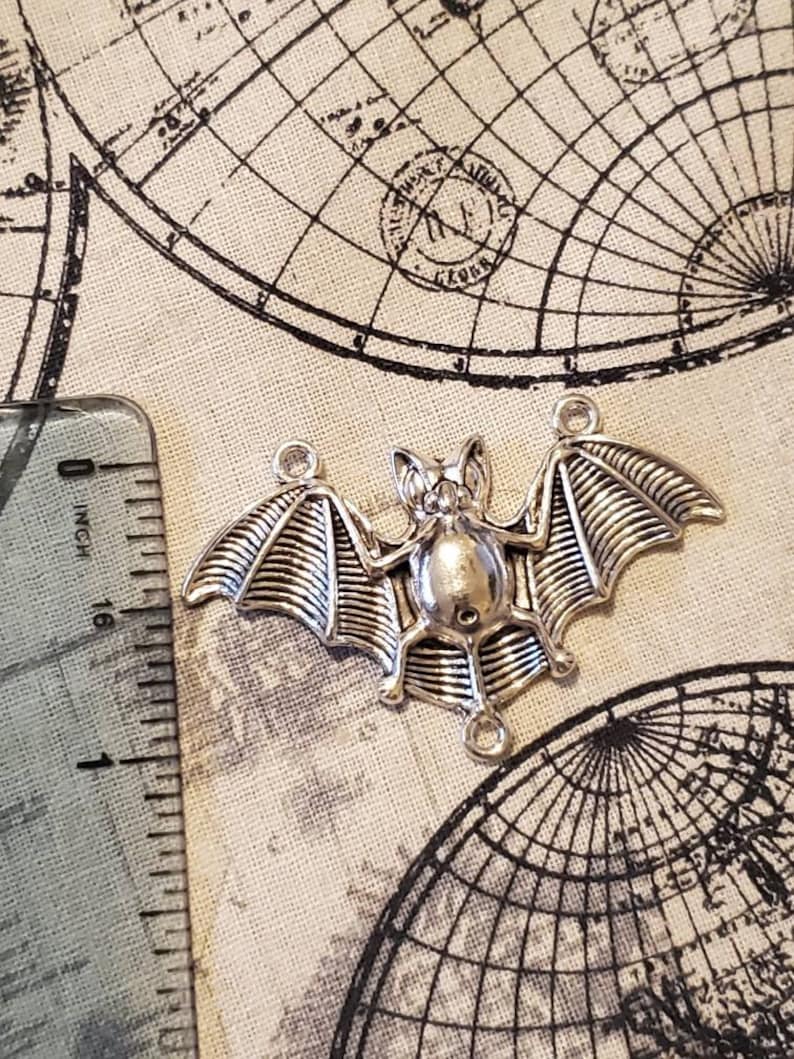 May include: Silver-toned metal bat charm with detailed wings and a hanging loop. The bat is approximately 1 inch in length.