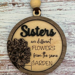 Puede incluir: Un adorno circular de madera con las palabras "Sisters are different FLOWERS from the same GARDEN" en letras negras. Un diseño floral está a la izquierda. El adorno cuelga de un cordón marrón y una cuenta de madera.