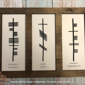 May include: A wooden framed wall art piece with three panels. Each panel features a white background with black Celtic symbols and text. The text reads "SLAINTE HEALTH", "GRA LOVE", and "SONAS HAPPINESS".