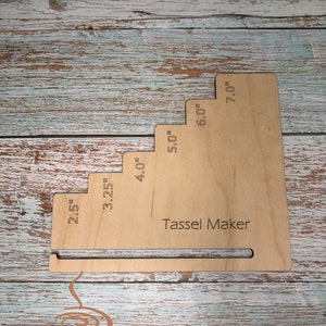 May include: A wooden Tassel Maker with measurements from 6.35 cm to 17.78 cm etched on the surface. The tool has a stepped design and a slot at the base. The text "Tassel Maker" is visible. The background is a weathered, light blue and brown wooden surface.