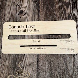 Puede incluir: Un letrero de madera con el texto "Canada Post Lettermail Slot Size" y las dimensiones para las ranuras de correo de tamaño grande y estándar. El letrero está hecho de madera clara y tiene un fondo marrón.