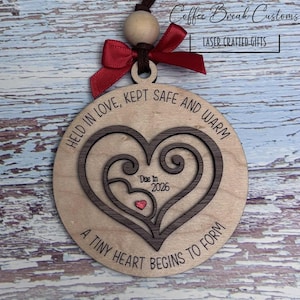 May include: A wooden heart-shaped ornament with a red bow and a brown string. The ornament features the text "HELD IN LOVE, KEPT SAFE AND WARM" and "A TINY HEART BEGINS TO FORM". It also says "Due in 2026".