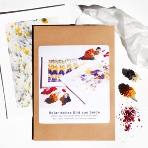 Könnte beinhalten: Eine braune Papierverpackung mit einem weißen Etikett mit dem Text "Botanisches Bild aus Seide". Im Inneren befinden sich Kunstdrucke mit farbenfrohen botanischen Designs. Getrocknete Blumen und Blütenblätter sind um die Verpackung herum verstreut, mit einem weißen Tuch im Hintergrund.