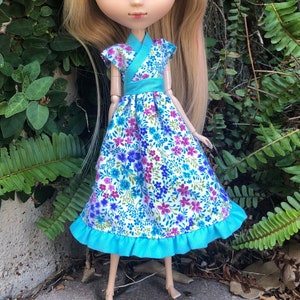 May include: A Pullip doll with blonde hair and green highlights, wearing a floral print dress with blue accents. The doll has large green eyes and is posed outdoors against a backdrop of green foliage.