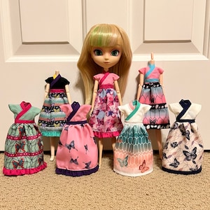 May include: Seven dolls wearing different colorful dresses with butterfly patterns. The dolls are all wearing different styles of dresses, some with long sleeves and some with short sleeves. The dresses are all made of different fabrics and have different embellishments.