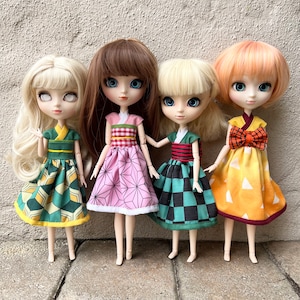 May include: Four dolls with long blonde, brown, and orange hair wearing colorful dresses. The dolls have large blue eyes and are posed standing on a gray textured surface.