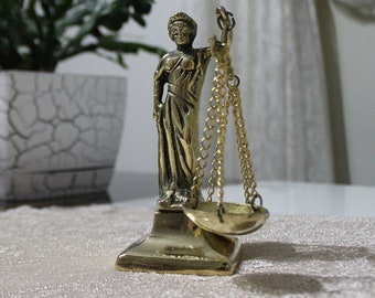 May include: A gold-colored statue of a woman holding a scale, symbolizing justice. The statue is on a white table with a white pot with a green plant in the background.