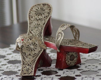 May include: A pair of ornate, silver filigree sandals with red platforms. The sandals are displayed on a dark wooden table with a white lace runner. The intricate metalwork creates a delicate, open design.