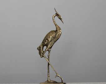 May include: A bronze-colored metal sculpture of a crane in a standing pose. The bird has a long, curved neck, a pointed beak, and detailed feathering. The crane stands on thin legs, with one leg slightly raised, and is mounted on a small, round base.