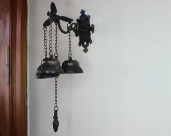 May include: A black metal door chime with three bells hanging from chains. The chime is mounted on a white wall.